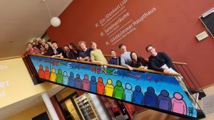 Auf diesem Bild sieht man eine bunt bemalte Treppe, auf der 13 Menschen stehen